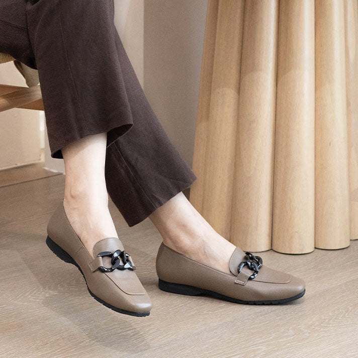 Person wearing brown loafers with decorative buckles on a wooden floor.